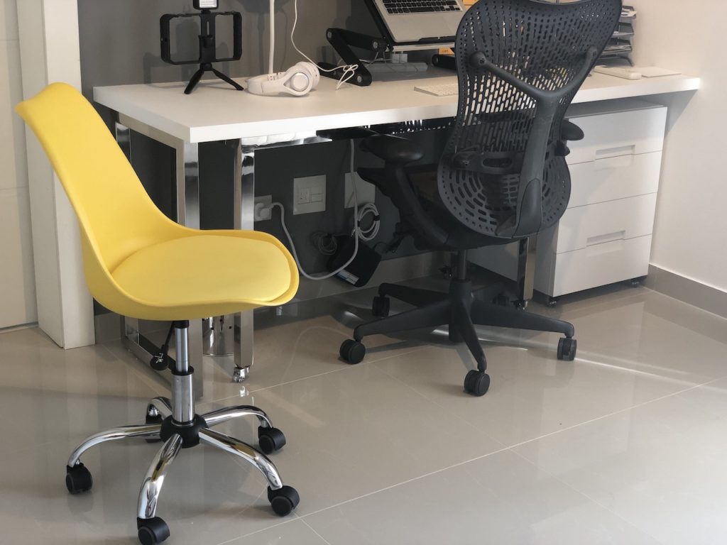 Home office com mesa de vidro recolhida na mesa fixa com cadeiras cinza e amarela giratórias.